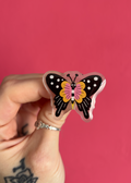 A hand with a silver ring holds a butterfly-shaped acrylic pin against a solid pink background. The pin features a black and white spotted wing pattern, a pink and yellow striped center, and small cartoon eyes.
