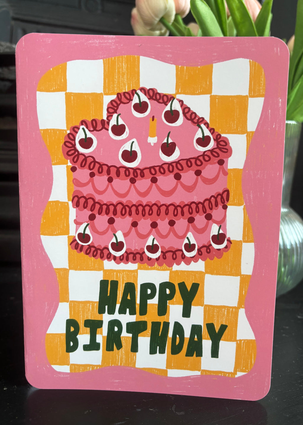 A pink 'Happy Birthday' greeting card featuring an illustration of a cherry-topped cake on a yellow and white checkered background, displayed on a dark mantelpiece in front of pink tulips.