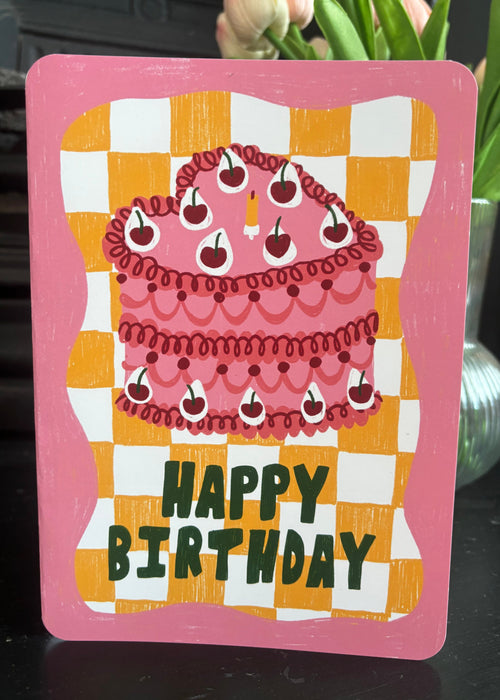 A pink 'Happy Birthday' greeting card featuring an illustration of a cherry-topped cake on a yellow and white checkered background, displayed on a dark mantelpiece in front of pink tulips.
