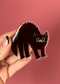 A hand with pink and black nail polish holds a die-cut acrylic pin featuring a stylized black cat with green eyes and a pink mouth, set against a solid pink background.