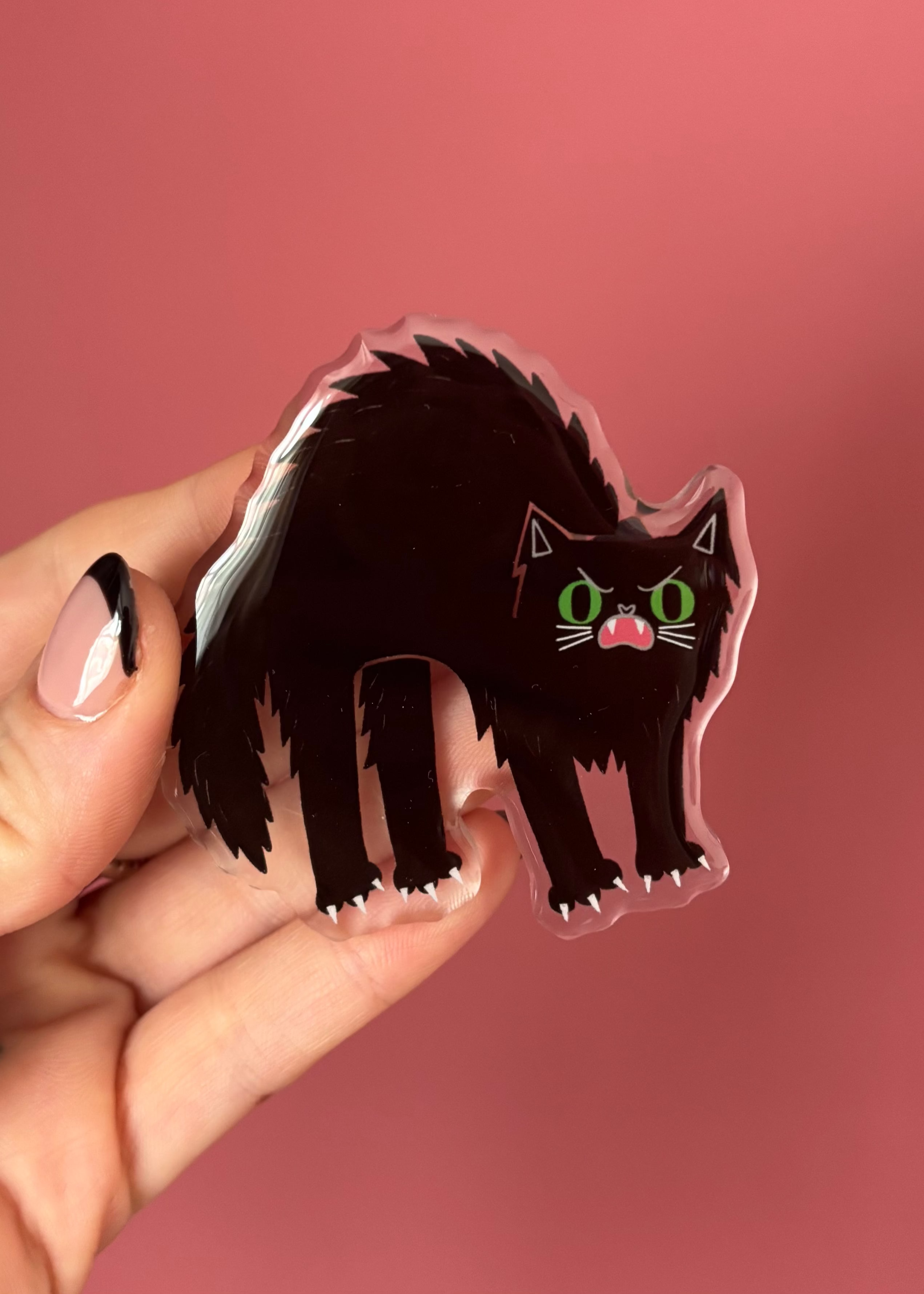 A hand with pink and black nail polish holds a die-cut acrylic pin featuring a stylized black cat with green eyes and a pink mouth, set against a solid pink background.