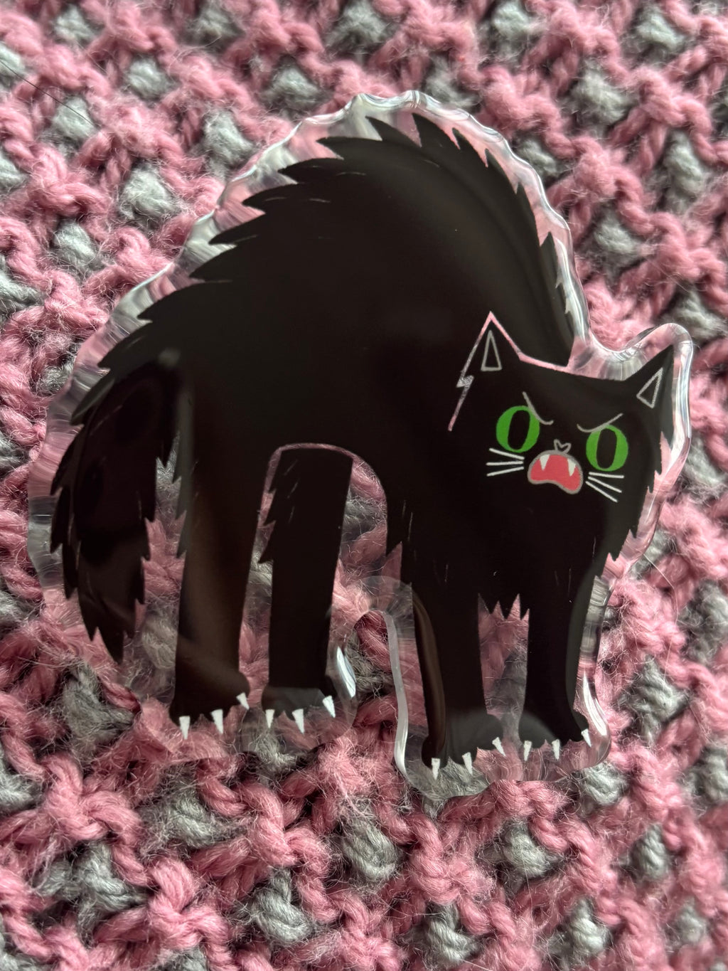 A black cat-shaped brooch with green eyes and a pink mouth, displayed on a pink and gray knitted textured background.