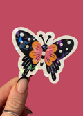 A hand with manicured nails holds a die cut butterfly sticker against a solid pink background. The butterfly features a black wing pattern with white dots on one side, orange wings on the other, and a pink and purple striped body.
