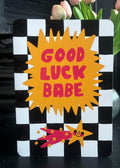A decorative sign with a black and white checkered border, a large yellow starburst in the center reading “GOOD LUCK BABE” in pink letters, and a small pink and yellow shooting star illustration at the bottom, shown standing on a dark surface with flowers behind it.