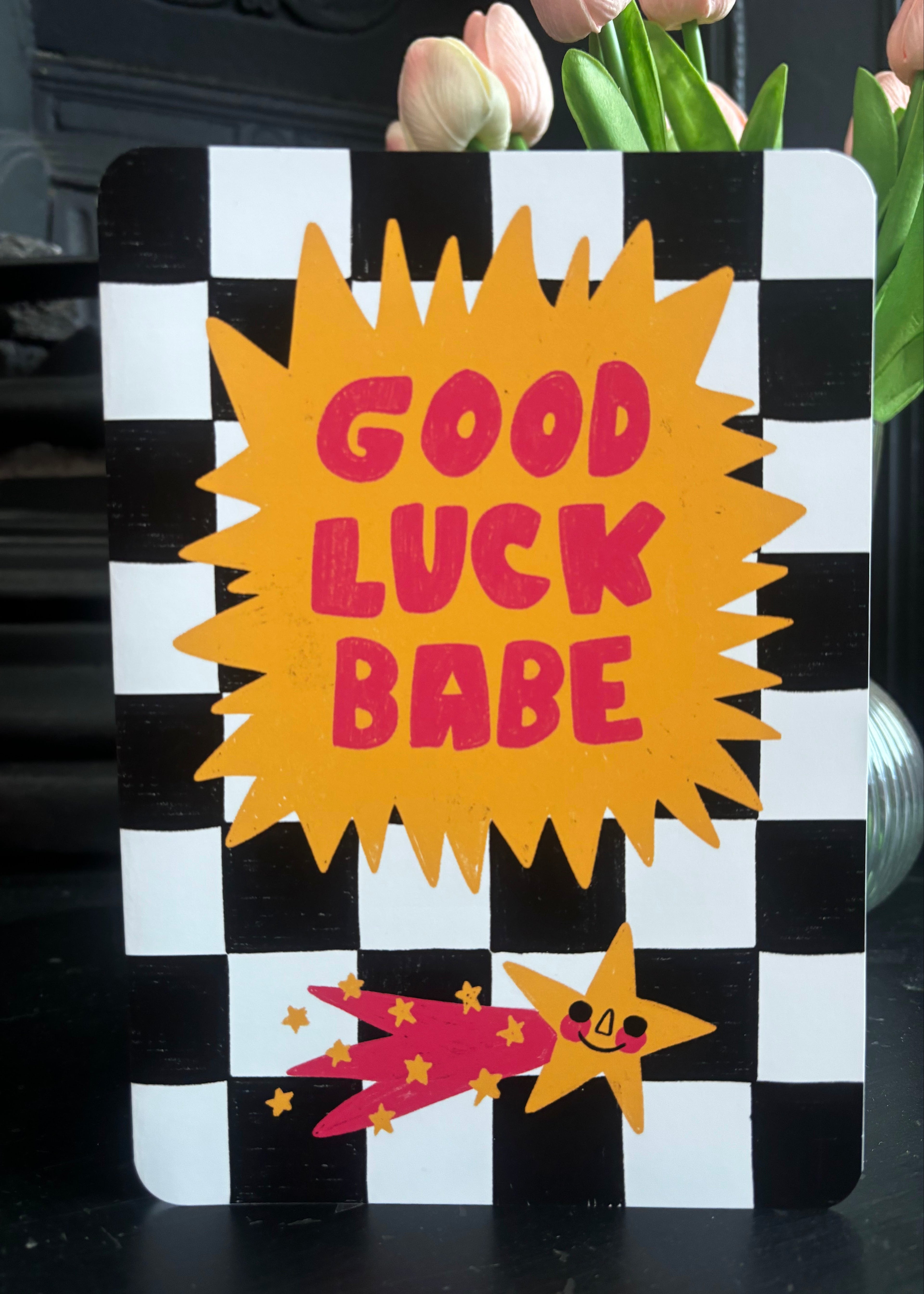 A decorative sign with a black and white checkered border, a large yellow starburst in the center reading “GOOD LUCK BABE” in pink letters, and a small pink and yellow shooting star illustration at the bottom, shown standing on a dark surface with flowers behind it.