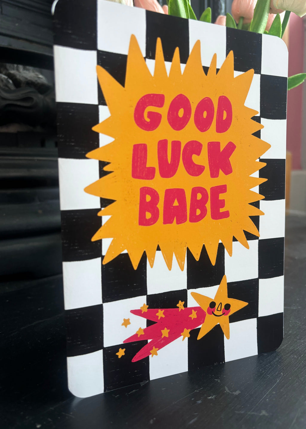 A rectangular decorative sign or card with a black-and-white checkerboard border, a large yellow starburst in the center reading “GOOD LUCK BABE” in pink letters, and a pink and yellow shooting-star graphic near the bottom, photographed on a dark tabletop with blurred background decor.