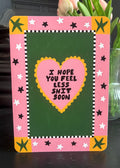 A square greeting card with a pink border, green center, and a yellow heart containing the text 'I HOPE YOU FEEL LESS SHIT SOON', displayed on a dark surface with tulips in the background.
