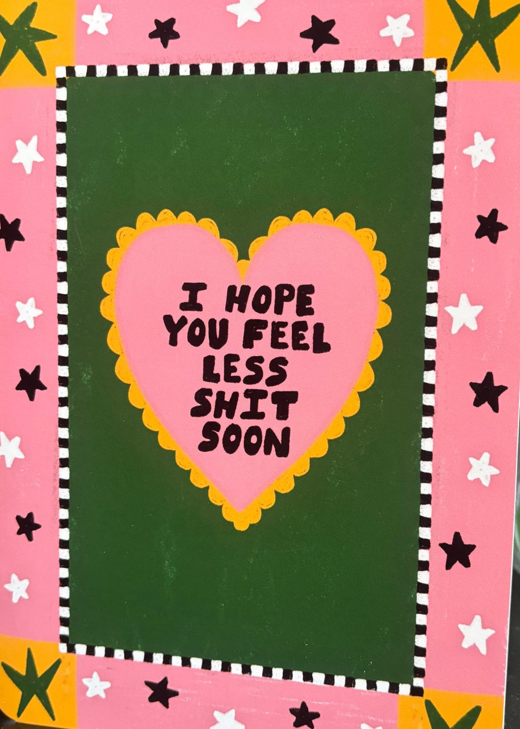 A greeting card with a pink border, yellow and white star accents, and a dark green center panel. A pink heart in the middle reads, 'I HOPE YOU FEEL LESS SHIT SOON' in black text.