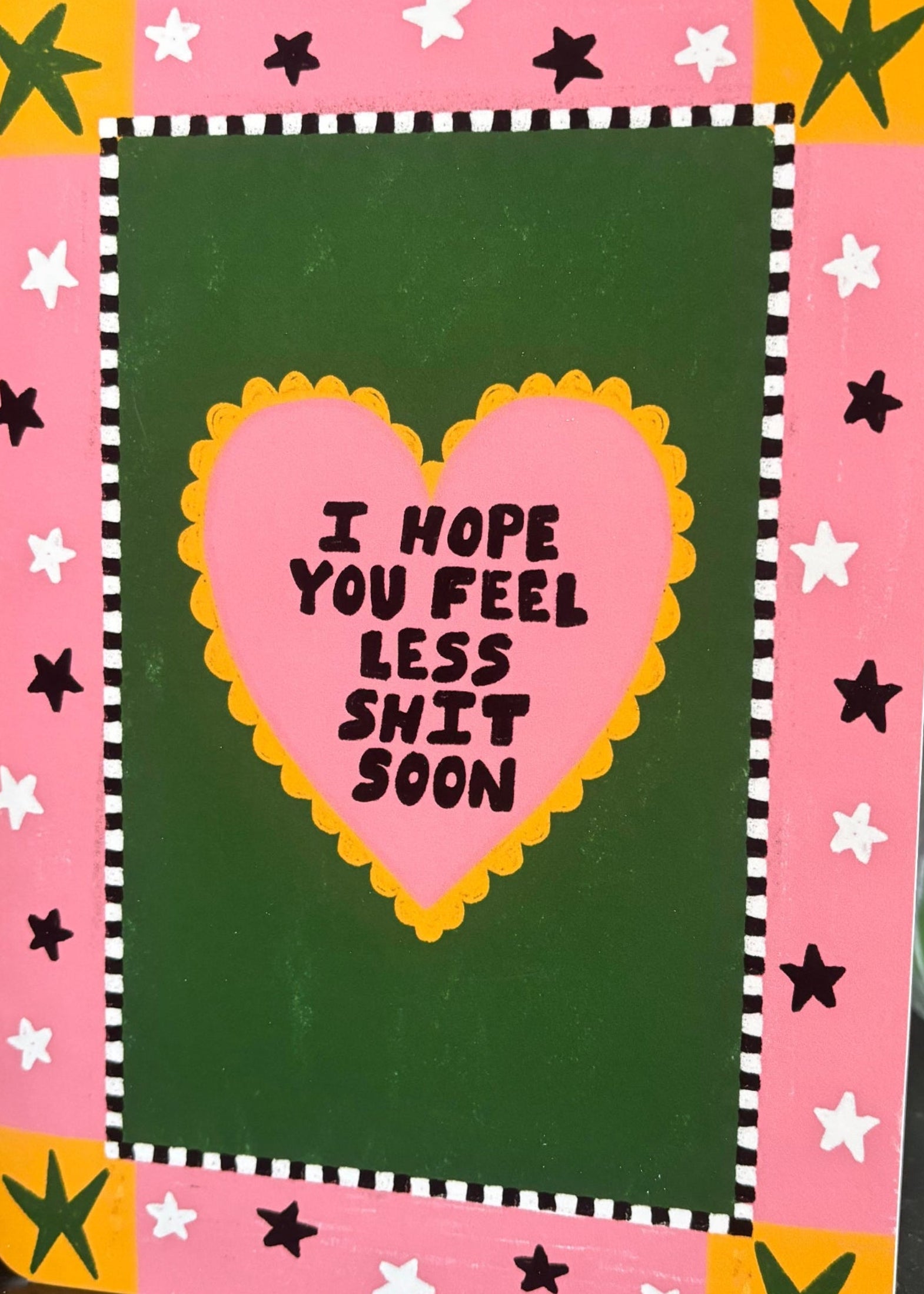 A greeting card with a pink border, yellow and white star accents, and a dark green center panel. A pink heart in the middle reads, 'I HOPE YOU FEEL LESS SHIT SOON' in black text.