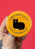 A hand with tattoos holds a round yellow button or badge featuring a black cat illustration and the text 'LIVE.LAUGH.