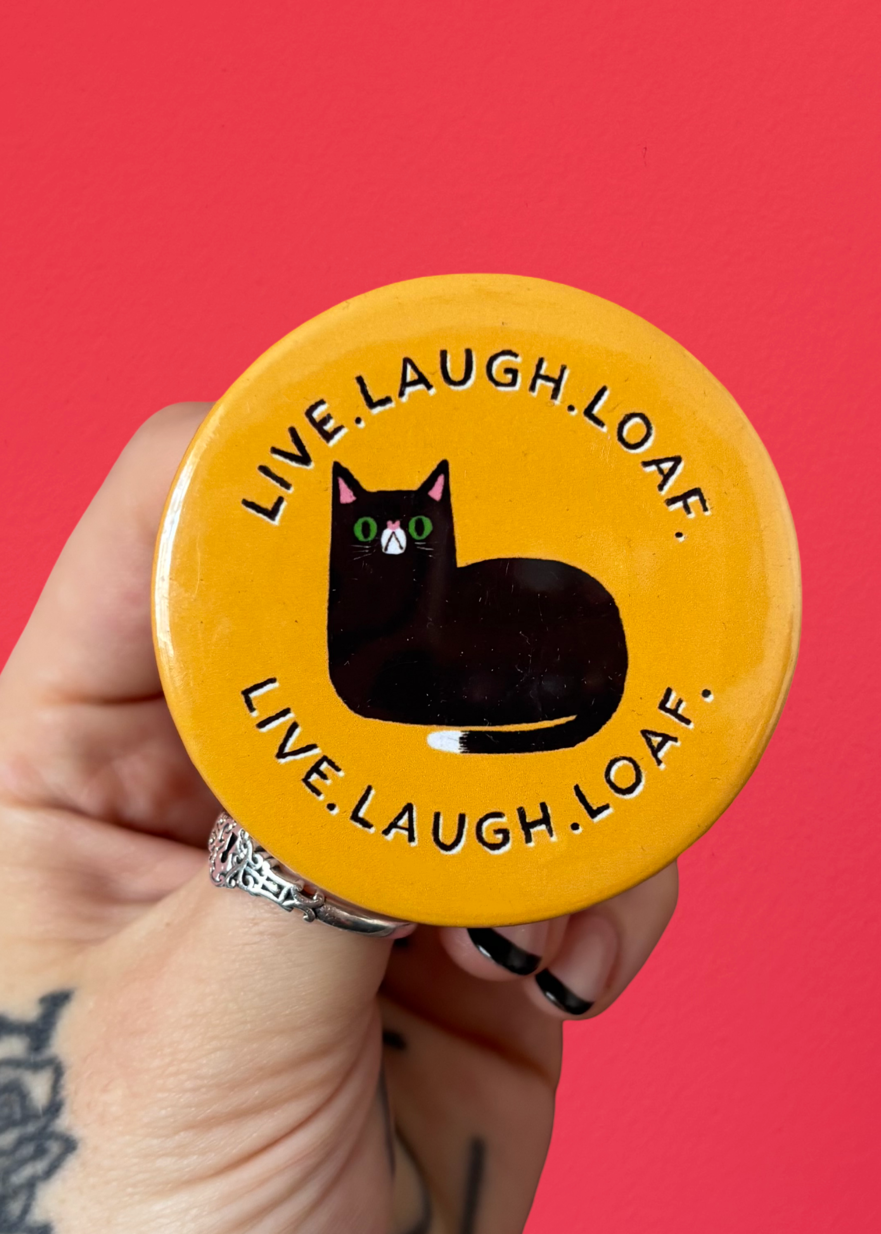 A hand with tattoos holds a round yellow button or badge featuring a black cat illustration and the text 'LIVE.LAUGH.