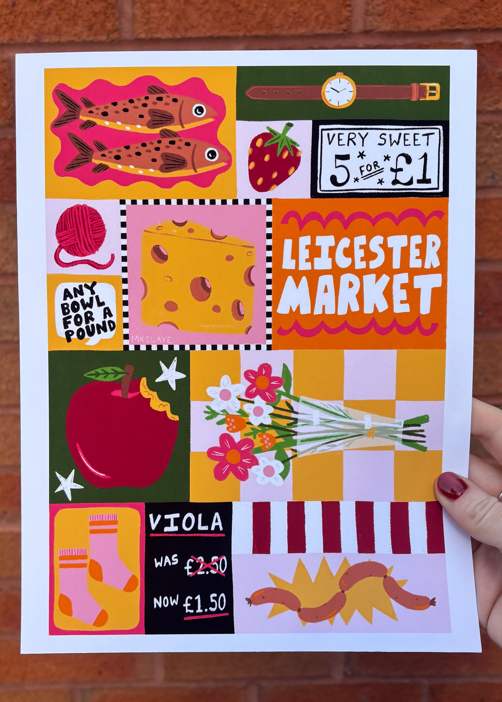 A hand holds a square art print featuring a colorful collage of Leicester Market-themed illustrations, including fish, a strawberry, cheese, an apple, flowers, socks, and a worm, against a red brick wall background.