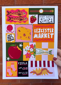 A hand holds a square art print featuring a colorful collage of Leicester Market-themed illustrations, including fish, a strawberry, cheese, an apple, flowers, socks, and a worm, against a red brick wall background.