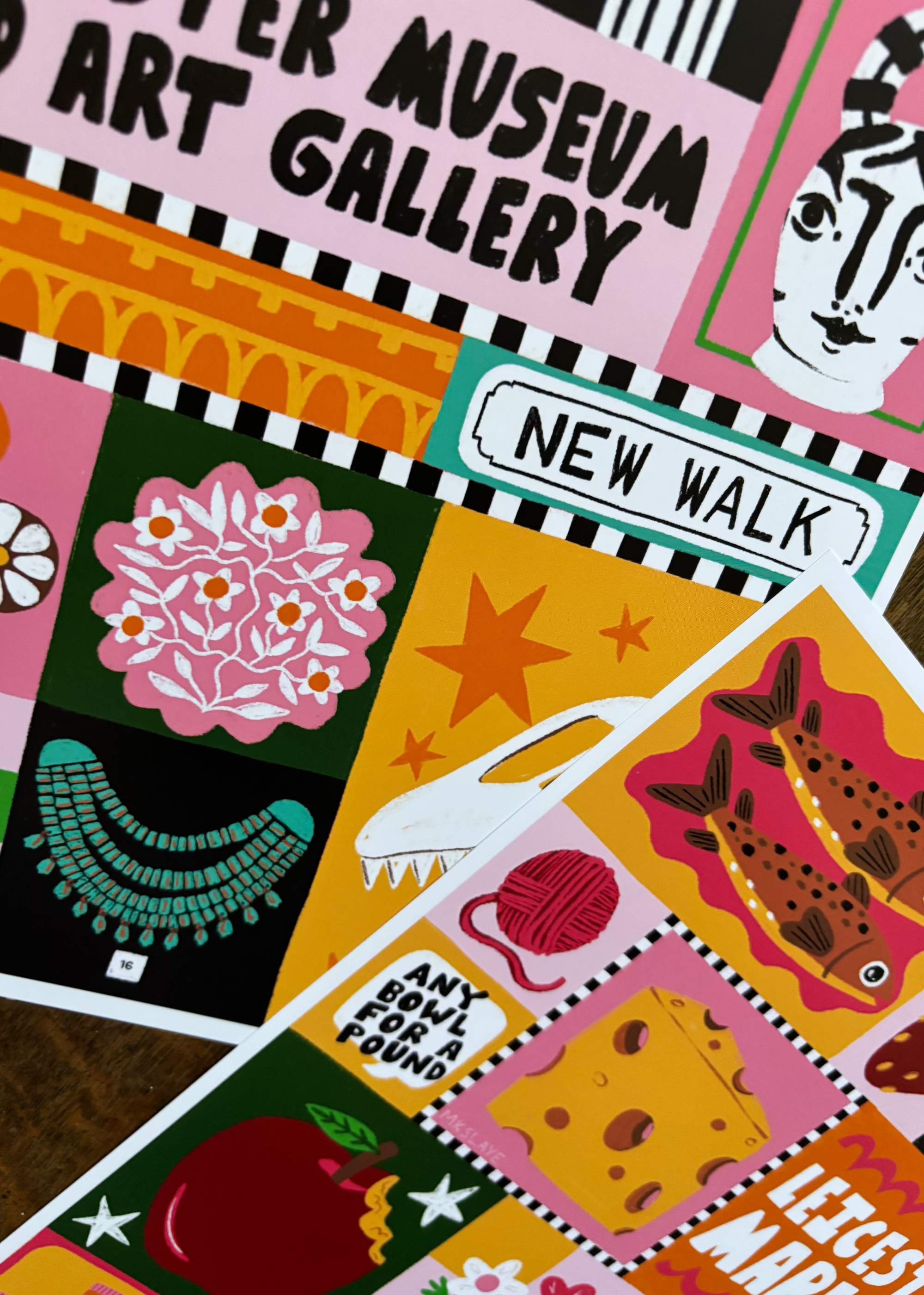 A close-up of overlapping illustrated postcards or printed cards featuring colorful abstract and whimsical designs, including text such as 'New Walk,' and 'Leicester Market,' on a wooden surface.