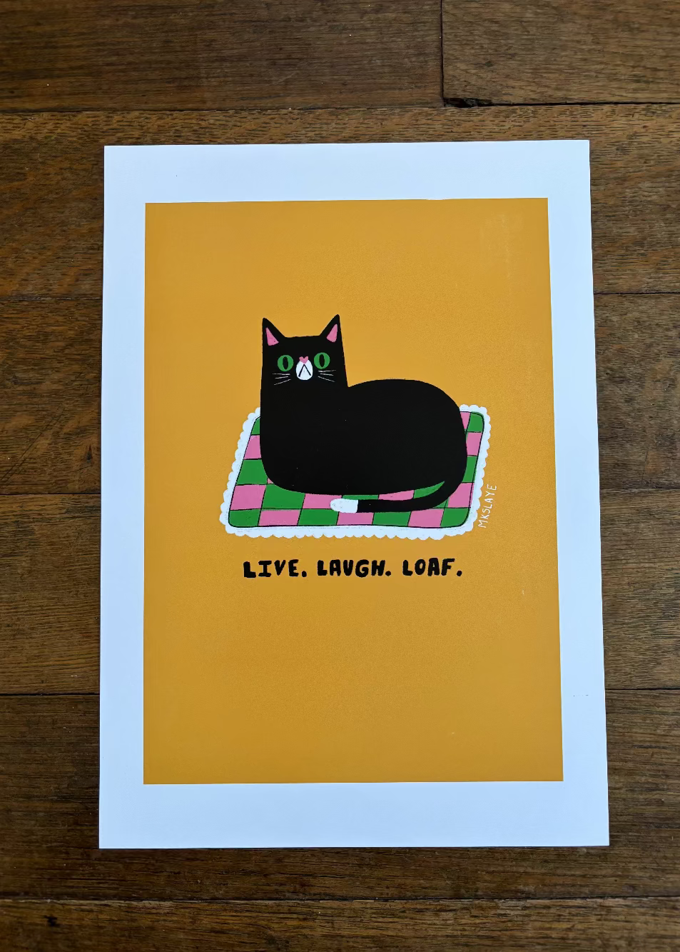 A square art print with a white border lies flat on a dark wood floor. The print features a black cat resting on a pink and green checkered blanket against a mustard yellow background, with the text 'LIVE, LAUGH, LOAF.