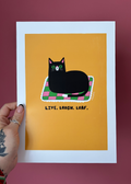 A hand holds a white-bordered art print featuring a black cat lying on a pink and green checkered blanket against a mustard yellow background, with the text 'LIVE, LAUGH, LOAF.' below.