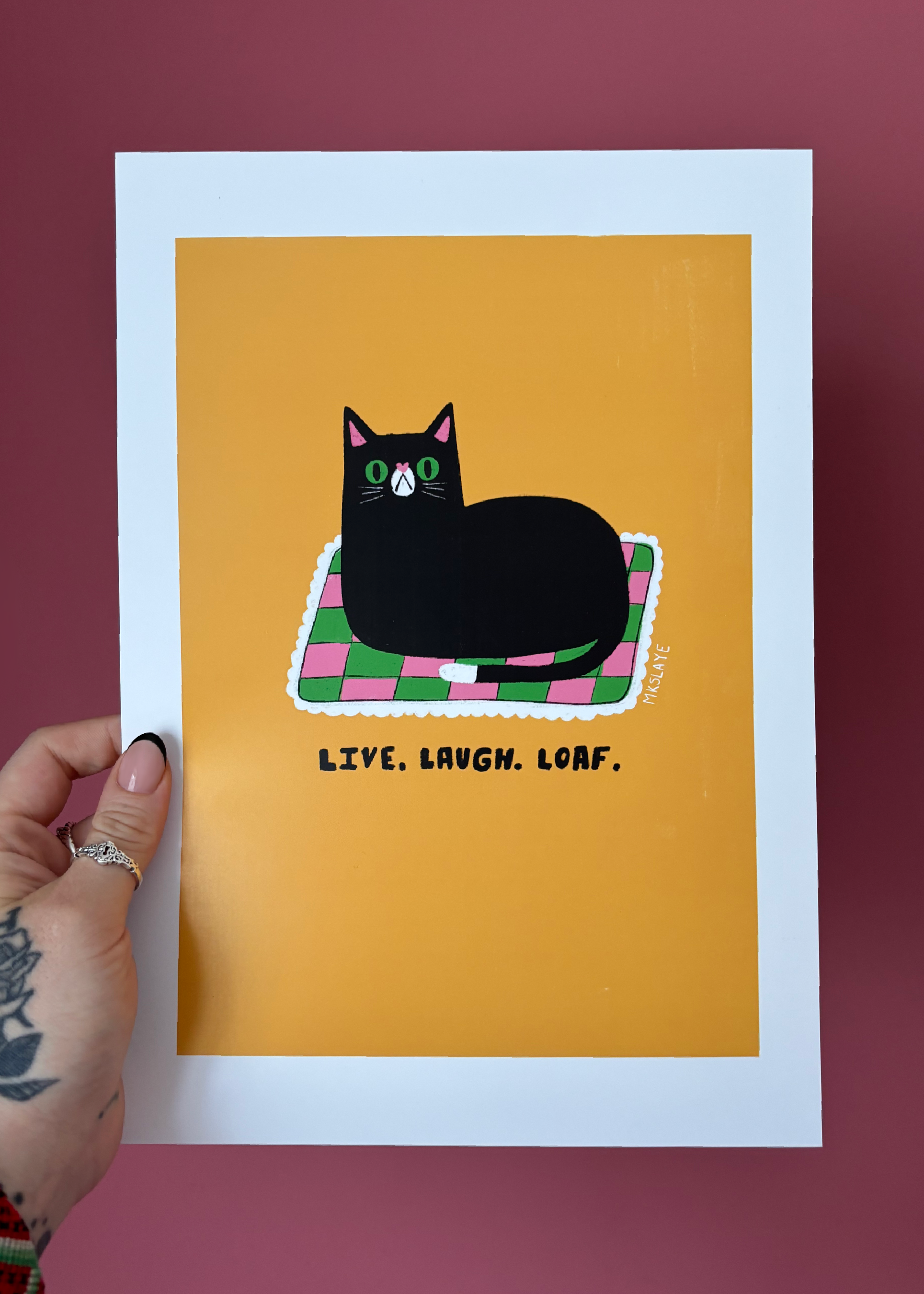 A hand holds a white-bordered art print featuring a black cat lying on a pink and green checkered blanket against a mustard yellow background, with the text 'LIVE, LAUGH, LOAF.' below.