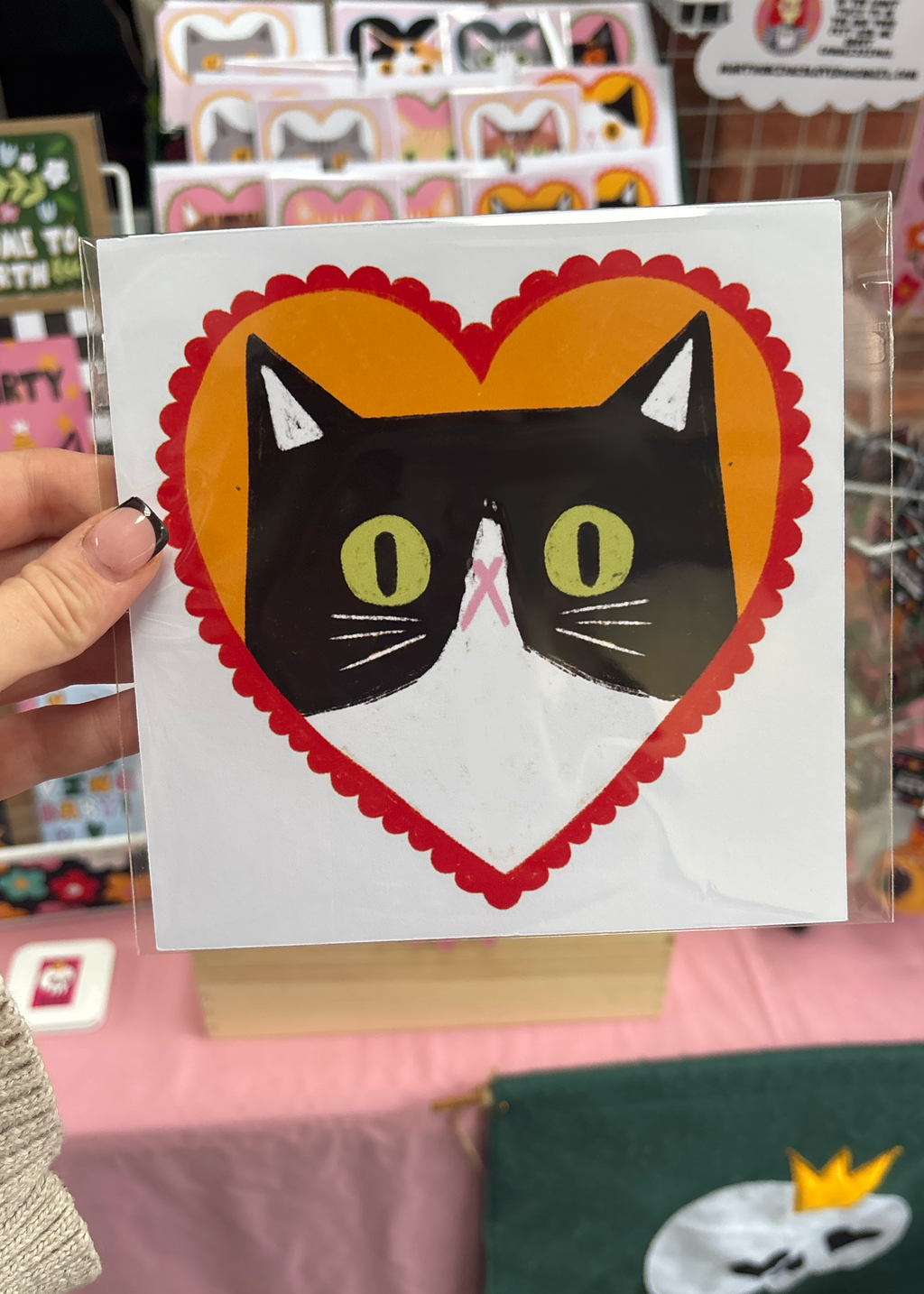 A hand holds a square white card featuring a stylized black-and-white cat face inside a large orange and red scalloped heart. The card is photographed in a retail or craft shop setting with shelves and colorful items blurred in the background.