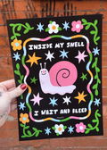 A hand holds a black rectangular art print featuring a pink snail and the text 'INSIDE MY SHELL I WAIT AND BLEED' against a brick wall background.