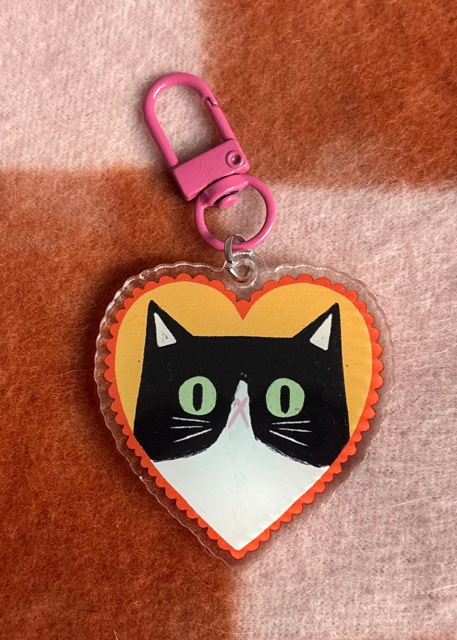 A heart-shaped acrylic keychain featuring a cartoon cat face with a black and white pattern, green eyes, and a pink nose, attached to a bright pink lobster clasp, set against a brown and beige checkered fabric background.