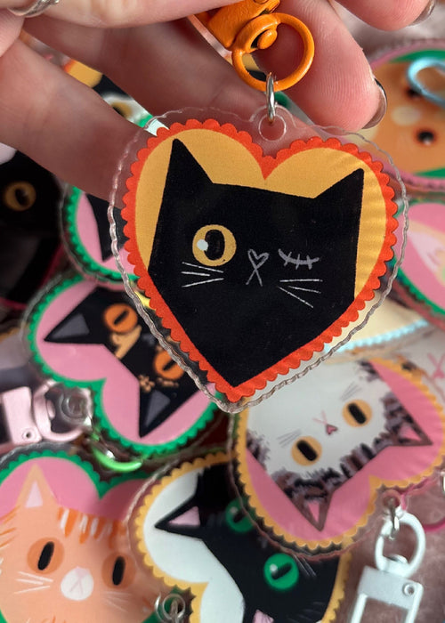 A hand holds a heart-shaped acrylic keychain featuring a black cat face with a yellow eye and white whiskers, set against a yellow background with orange scalloped edges. Multiple similar keychains are visible in the background.