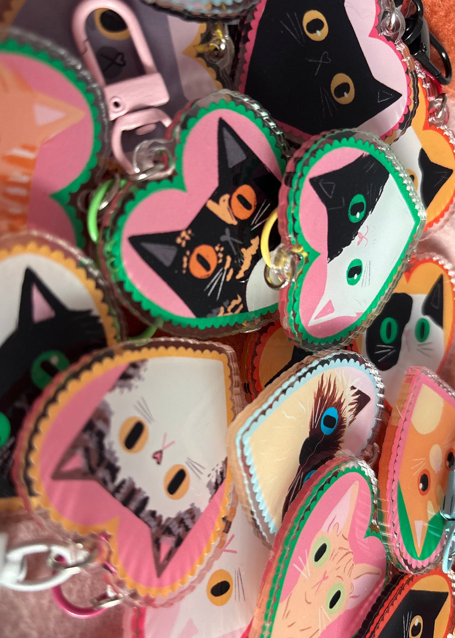 A close-up pile of heart-shaped acrylic keychains featuring different cat illustrations in pink, black, white, orange, and green.