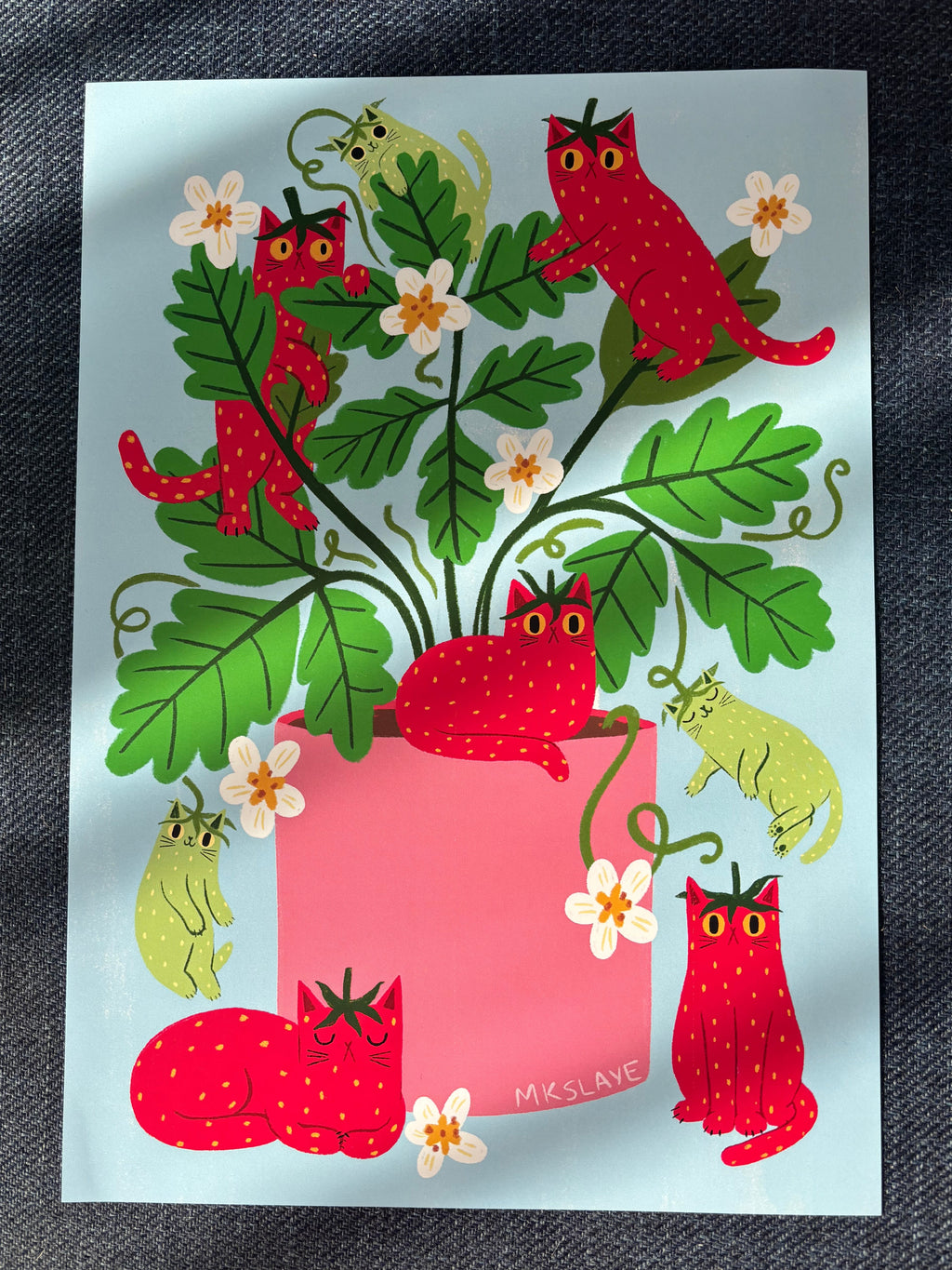An A4 art print featuring whimsical red cats shaped like strawberries, green leaves, and small white flowers on a pale blue background, placed on a dark textured fabric surface.