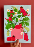 A hand holds a square art print featuring a whimsical illustration of red strawberry cats climbing and resting on a pink planter among green leaves and white flowers, set against a dark red background.