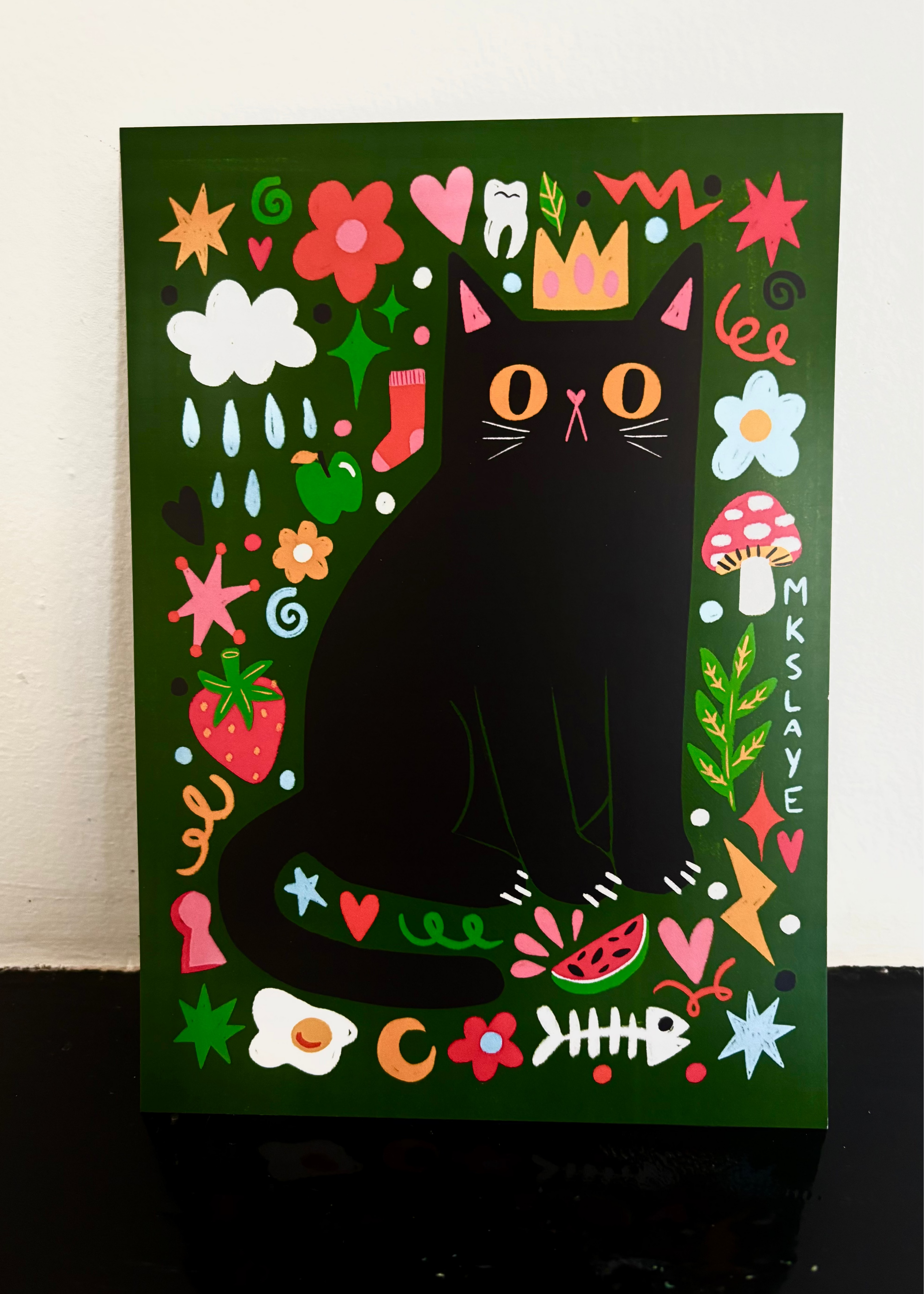 A square art print or poster featuring a whimsical illustration of a black cat with a gold crown, surrounded by colorful flowers, hearts, stars, mushrooms, and other playful motifs on a dark green background. The print is photographed leaning against a white wall on a dark surface.