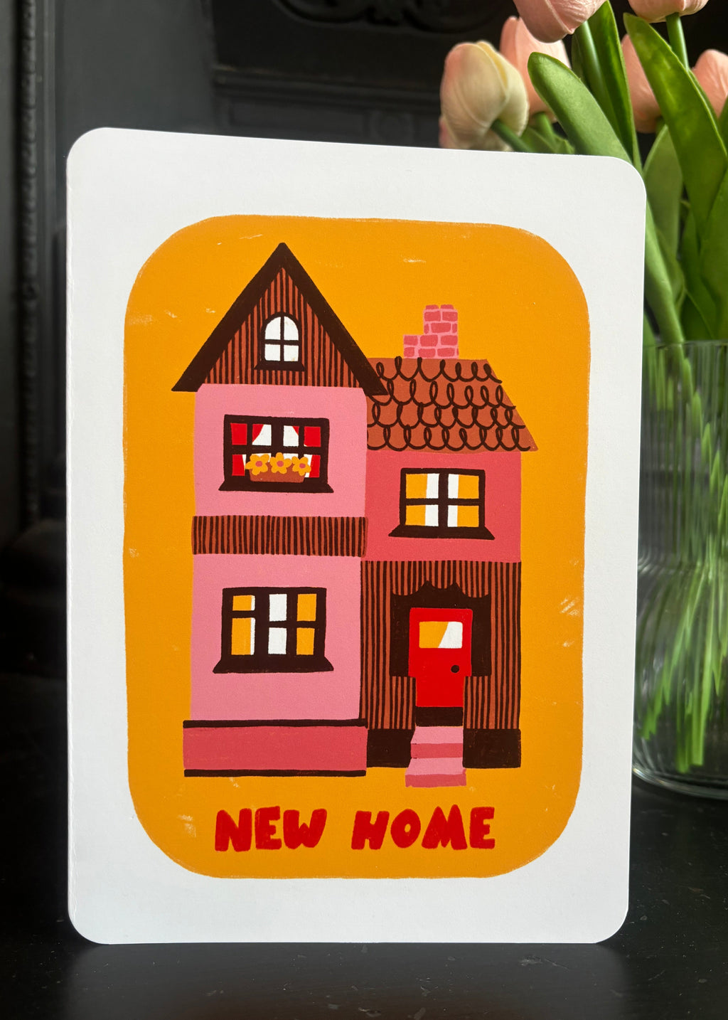 A square white greeting card with rounded corners featuring a colorful illustration of a pink house on an orange background, with the words 'NEW HOME' in red at the bottom. The card is photographed standing upright on a dark surface with pink tulips in a glass vase blurred in the background.