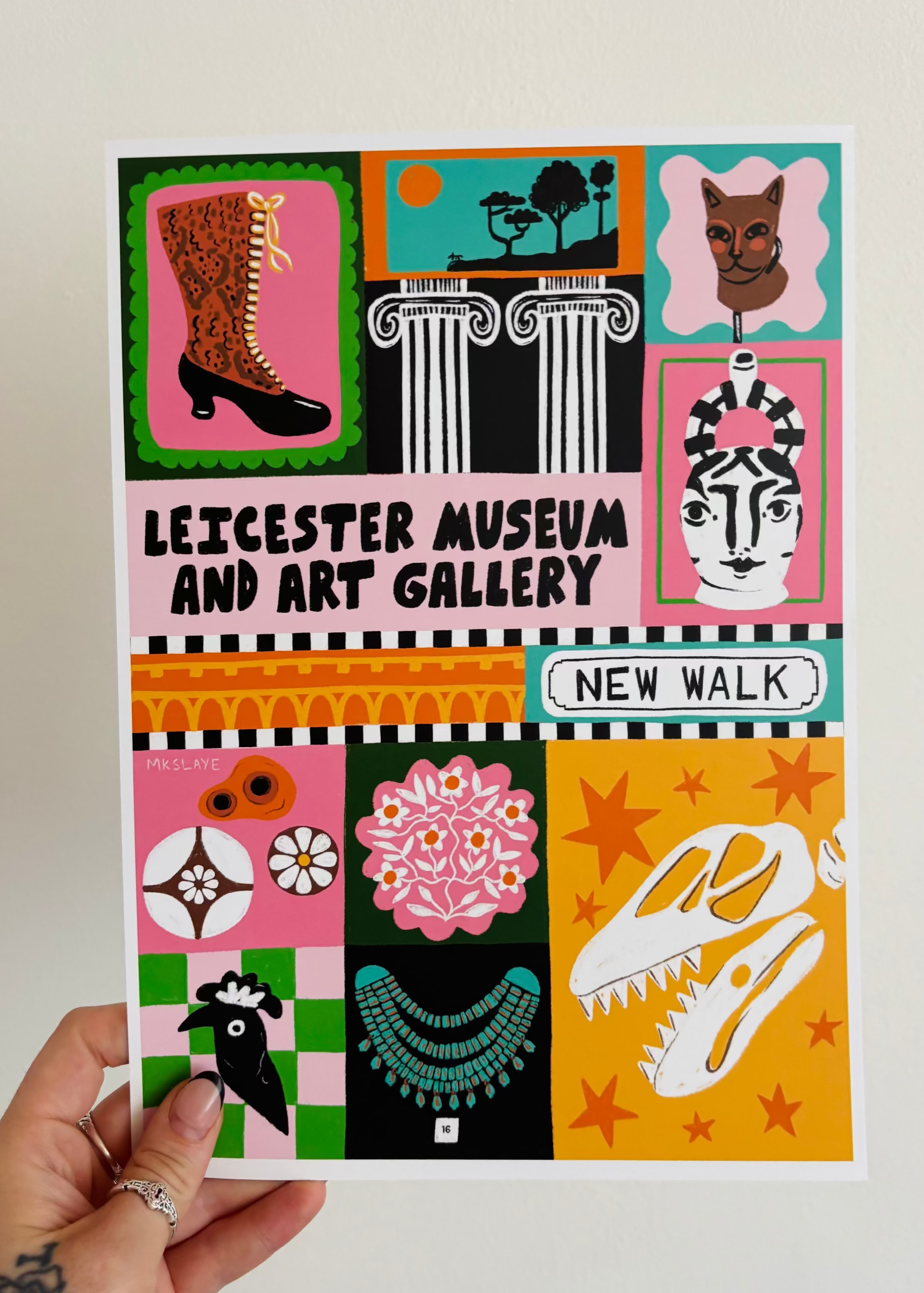 A hand holds a colorful illustrated poster for Leicester Museum and Art Gallery, New Walk, featuring a grid of whimsical drawings in pink, yellow, green, black, and white against a plain light background.