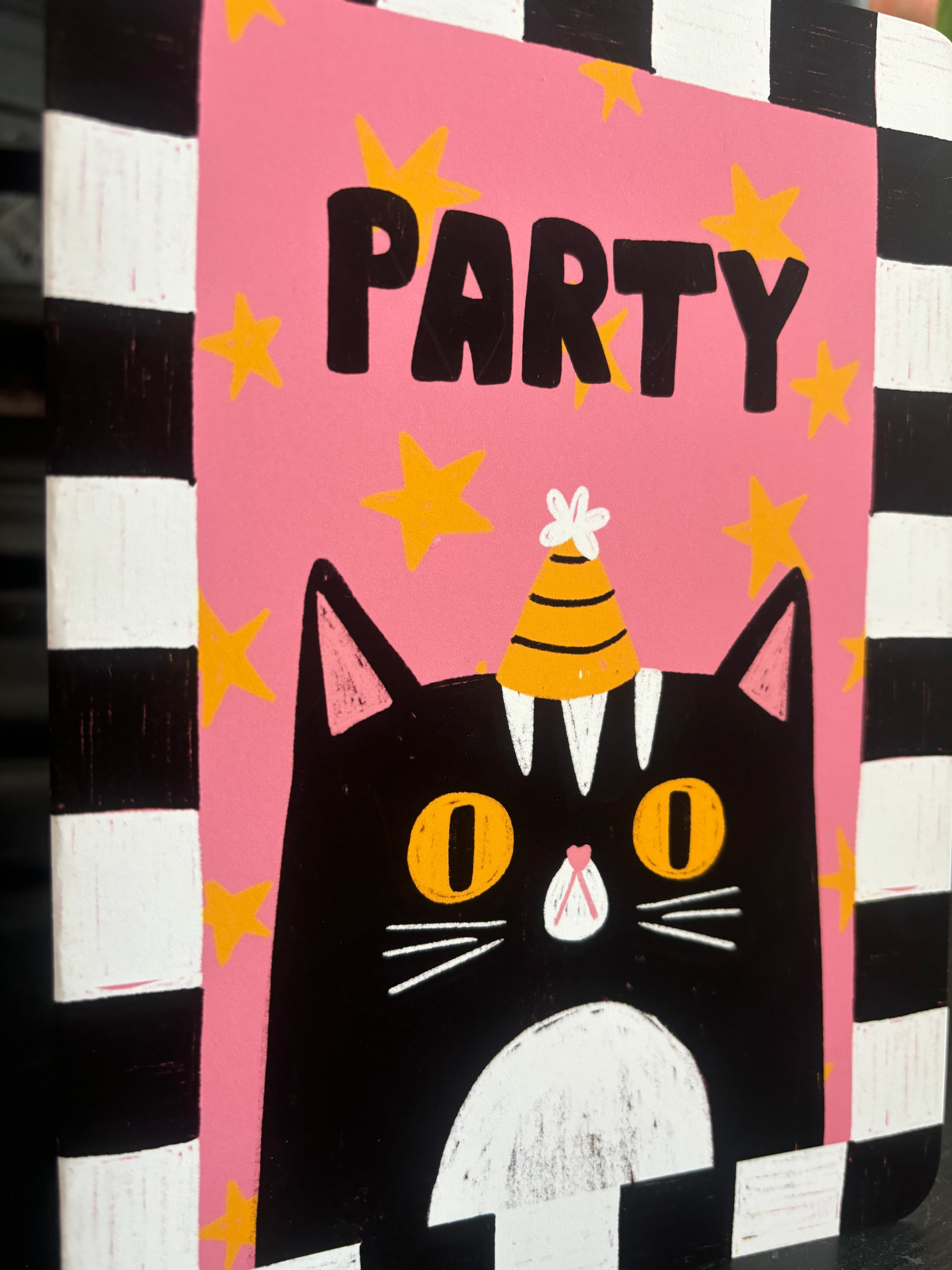 A square decorative sign or plaque featuring a stylized black cat with yellow eyes, a yellow party hat, and the word 'PARTY' in black letters on a pink background with yellow stars. The sign is photographed against a black-and-white striped backdrop.