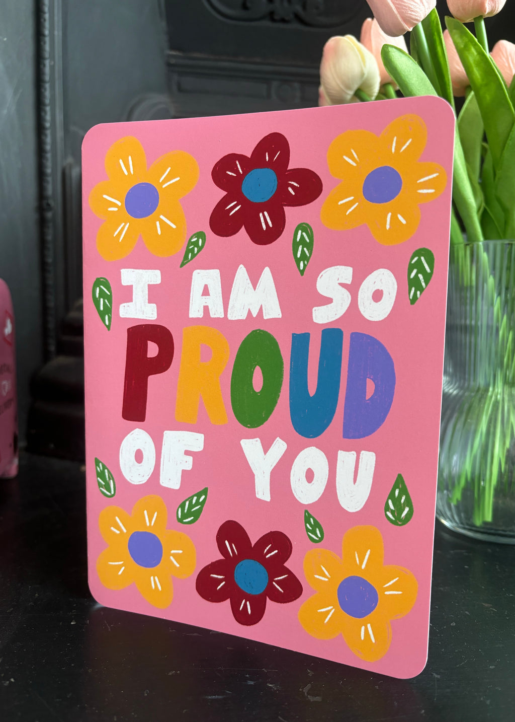 A pink greeting card with colorful hand-drawn flowers and the text 'I AM SO PROUD OF YOU' in multicolored letters, standing on a dark surface with tulips in the background.