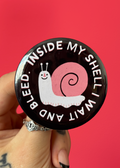 A hand with a silver ring holds a round black pinback button featuring a white and pink snail and the text 'INSIDE MY SHELL I WAIT AND BLEED.' against a solid red background.