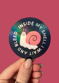 A hand holds a round sticker against a solid pink background. The sticker features a black background with a pink snail illustration and the text 'INSIDE MY SHELL I WAIT AND BLEED'.