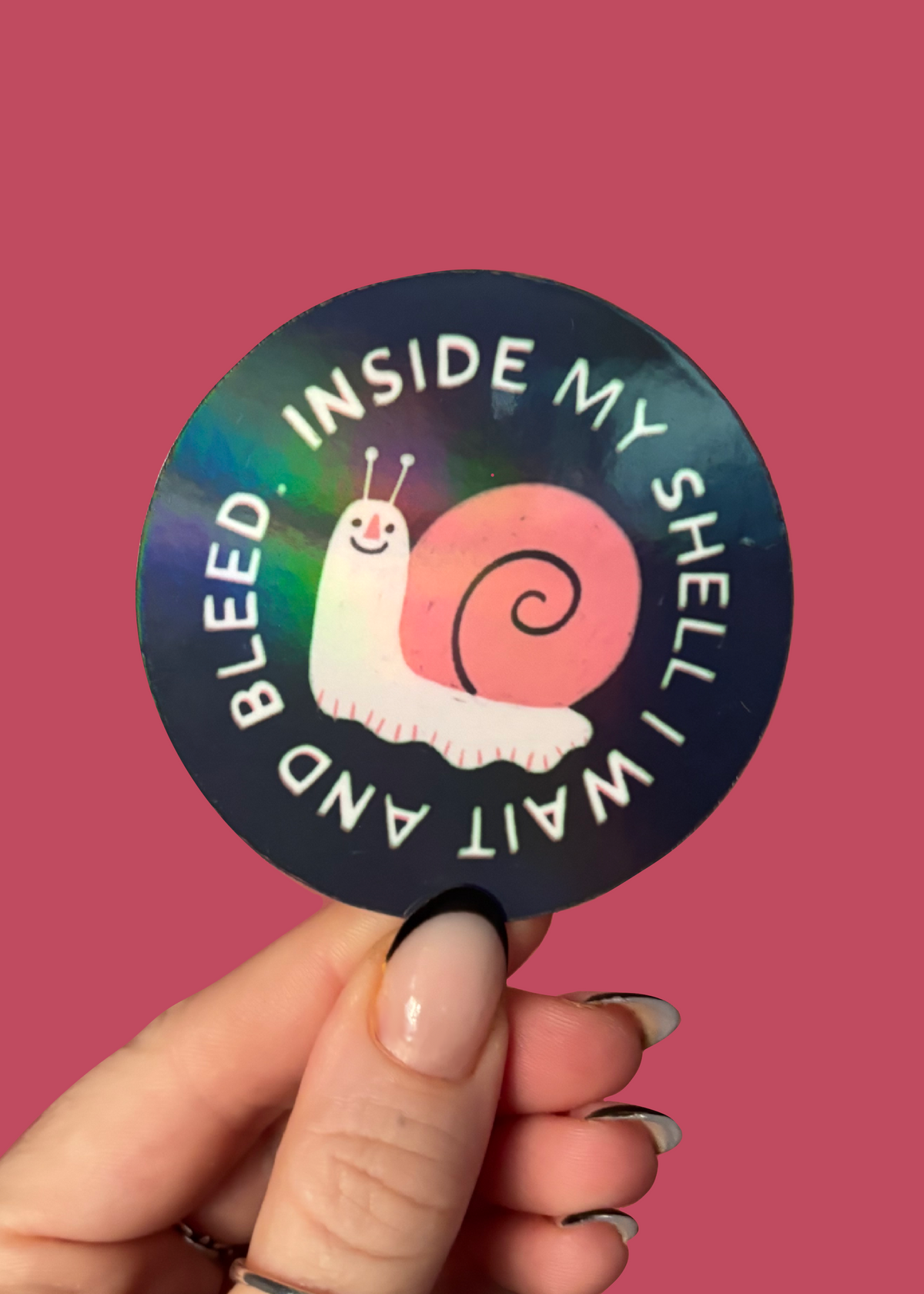 A hand holds a round sticker against a solid pink background. The sticker features a black background with a pink snail illustration and the text 'INSIDE MY SHELL I WAIT AND BLEED'.