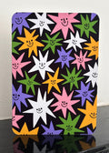 A rectangular black card or sign with rounded corners, featuring a colorful pattern of smiling starbursts in pink, purple, green, yellow, and white. The sign is displayed standing upright on a glossy black surface against a plain off-white wall.