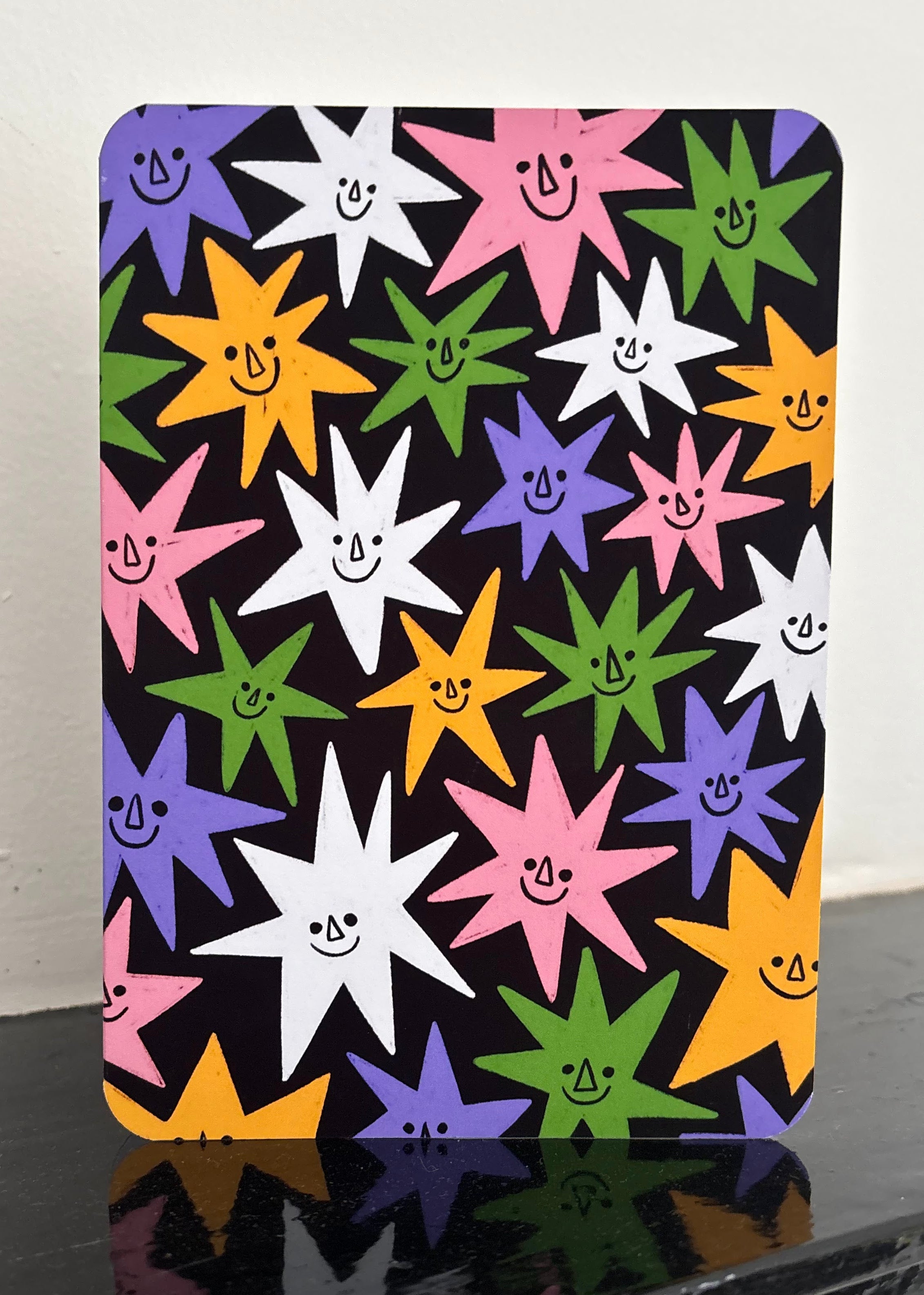 A rectangular black card or sign with rounded corners, featuring a colorful pattern of smiling starbursts in pink, purple, green, yellow, and white. The sign is displayed standing upright on a glossy black surface against a plain off-white wall.