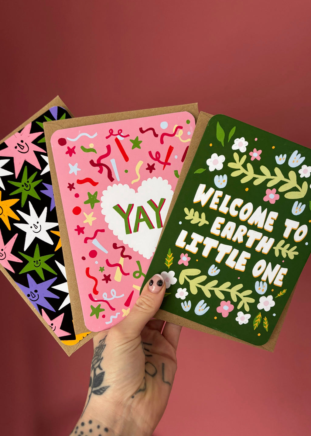 A hand with tattoos holds three colorful greeting cards against a dusty rose background. The front card is dark green with white text reading 'WELCOME TO EARTH LITTLE ONE' and white flowers with green leaves.