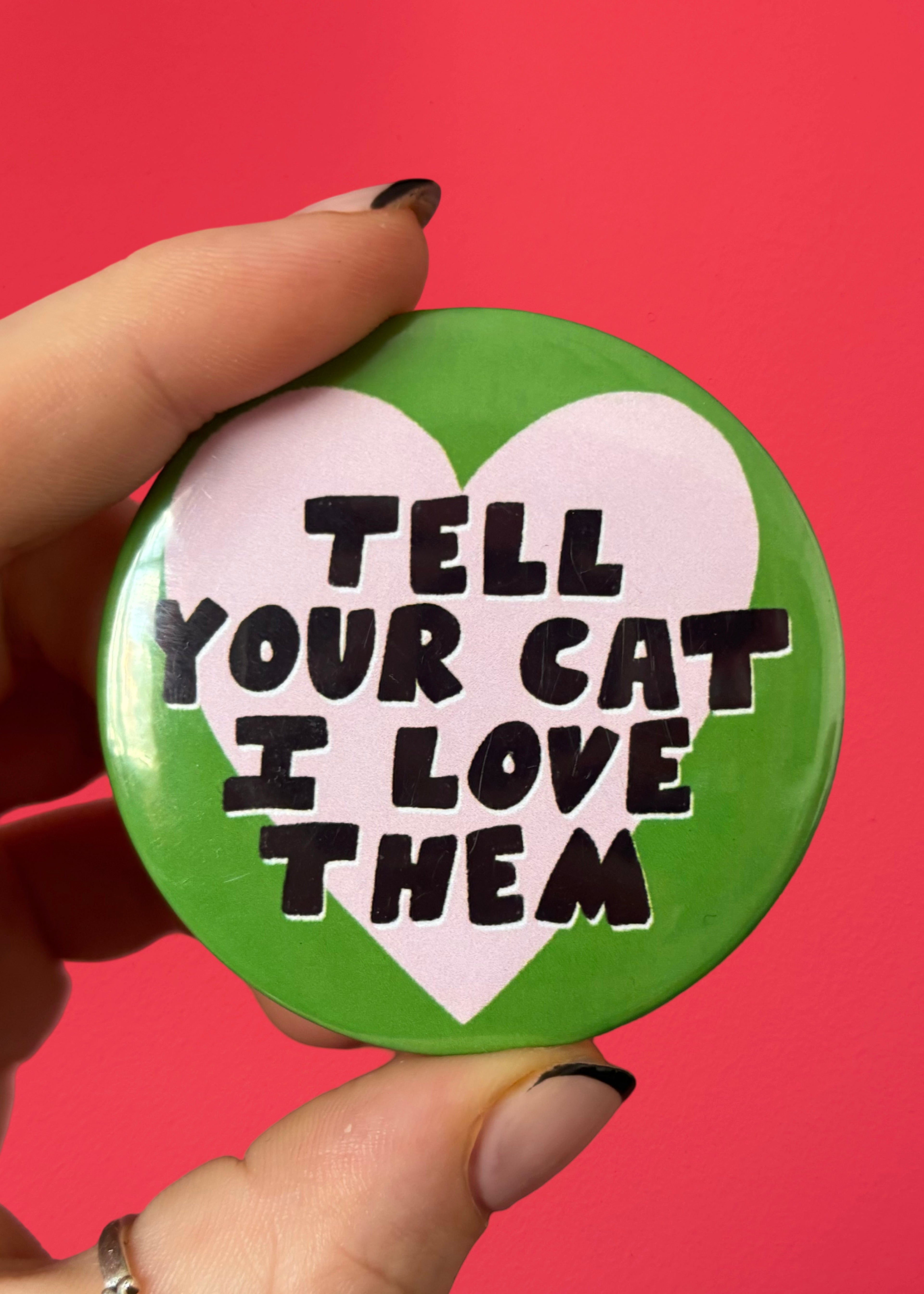 A hand holding a round pinback button with a green border, a white heart, and the text 'TELL YOUR CAT I LOVE THEM' in black letters, set against a solid red background.