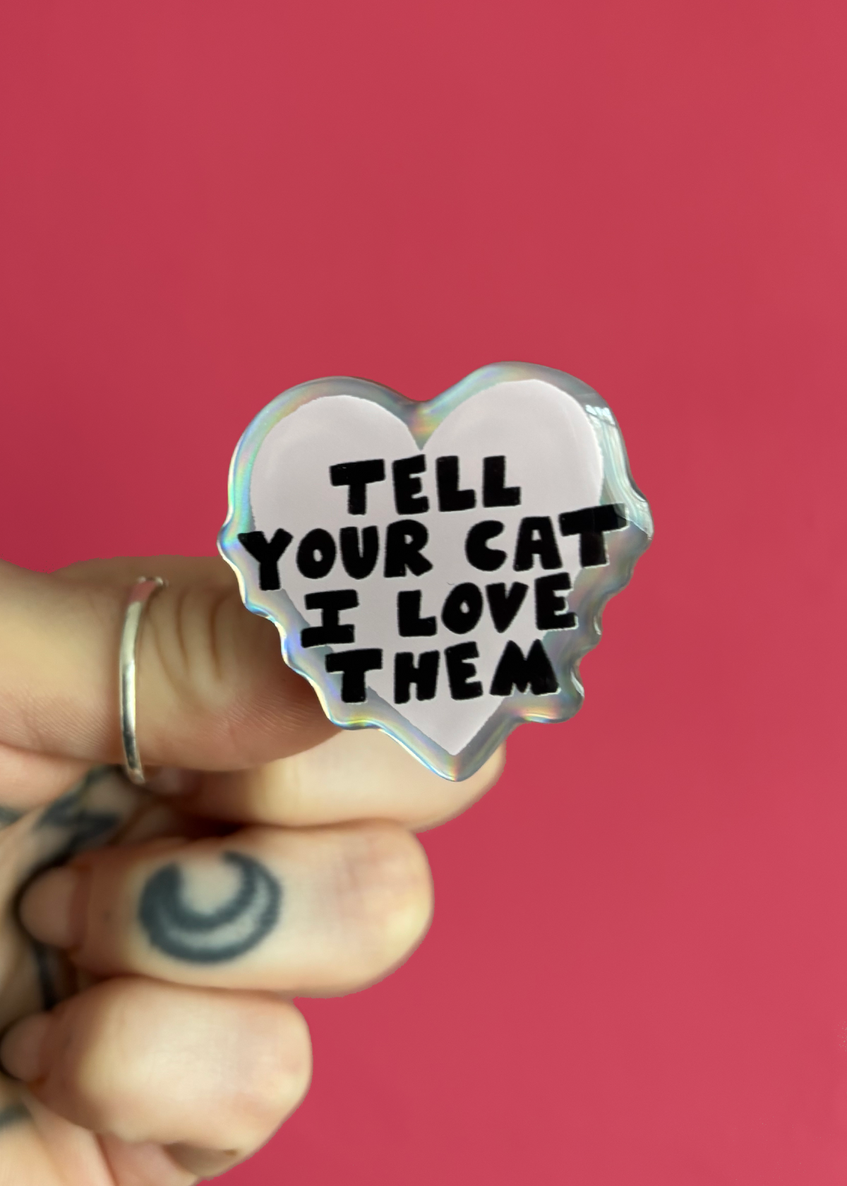 A hand with a silver ring and a crescent moon tattoo holds a heart-shaped holographic pin with the text 'TELL YOUR CAT I LOVE THEM' in black lettering against a solid pink background.
