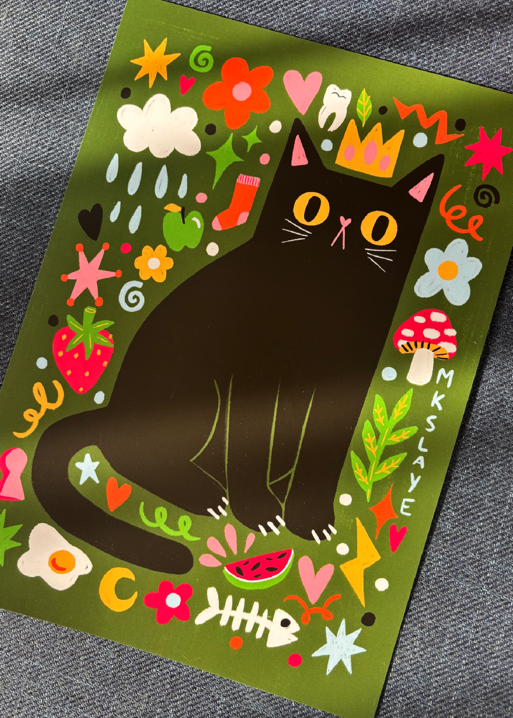 A square art print featuring a whimsical illustration of a black cat wearing a gold crown, surrounded by colorful flowers, mushrooms, stars, and food motifs on an olive green background, photographed on a gray textured fabric surface.