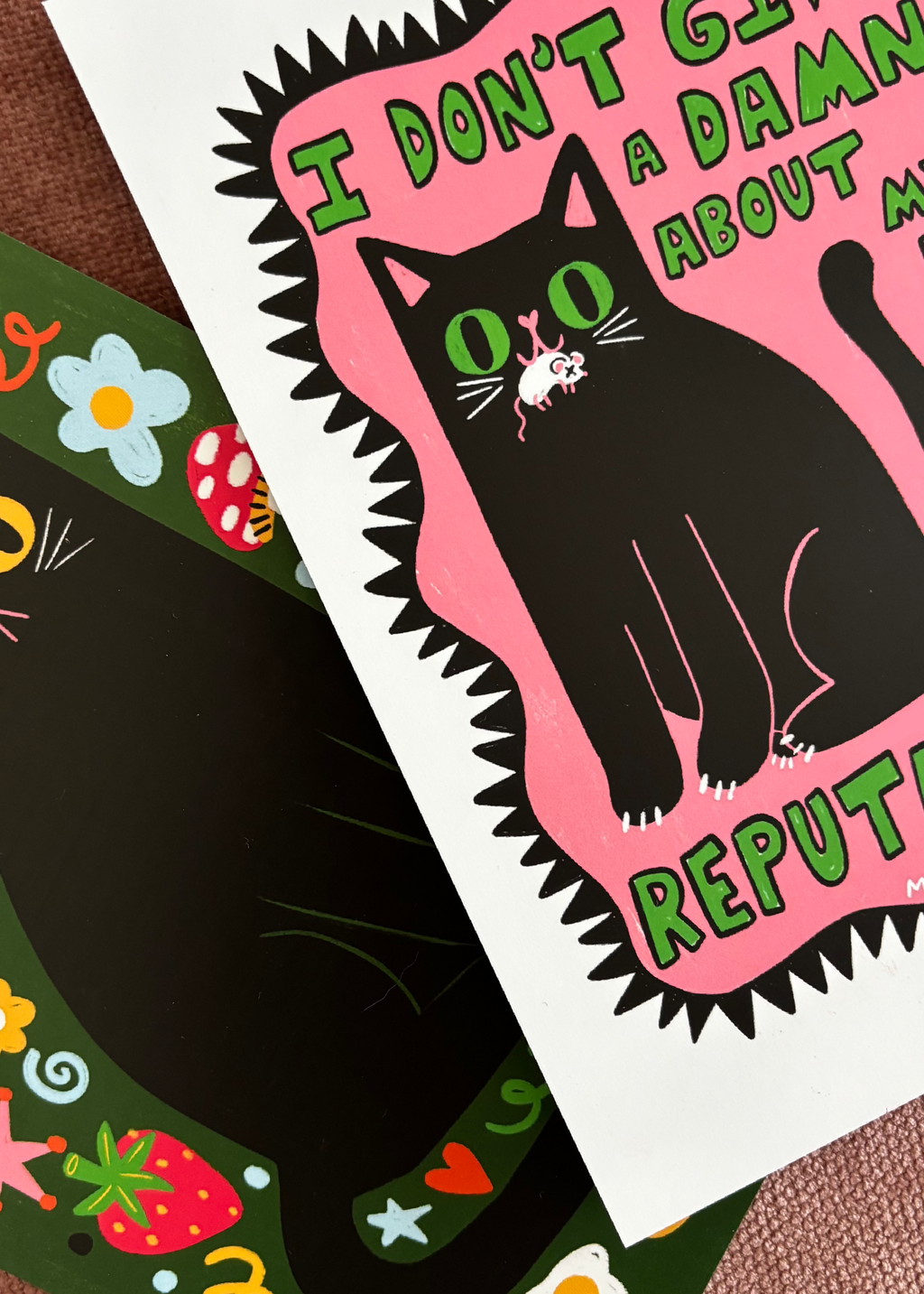 A close-up of a printed card or art print featuring a black cat on a pink shape with green text reading 'I DON'T GIVE A DAMN ABOUT MY BAD REPUTATION'