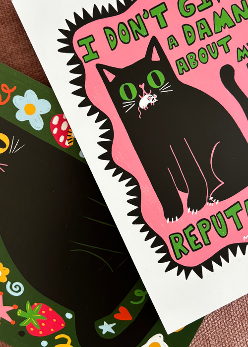 A close-up of a printed card or art print featuring a black cat on a pink shape with green text reading 'I DON'T GIVE A DAMN ABOUT MY BAD REPUTATION'