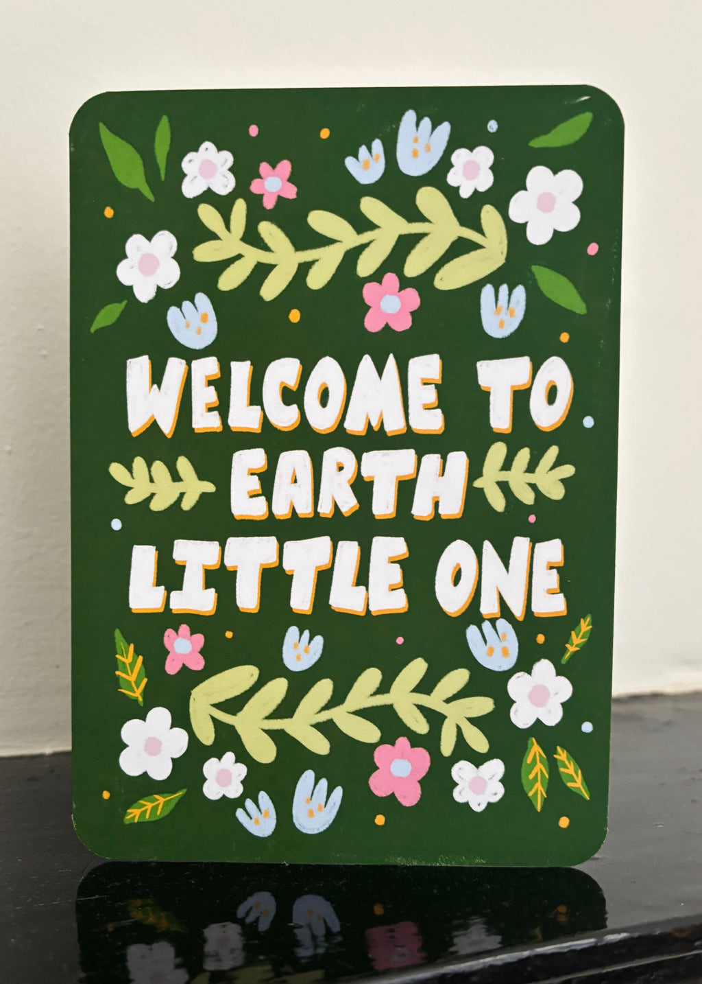 A rectangular dark green decorative card with rounded corners, featuring the phrase “WELCOME TO EARTH LITTLE ONE” in large white letters with yellow outlines, surrounded by hand-painted style flowers, leaves, and small handprints. The sign is photographed leaning on a glossy black surface against a light off-white wall.