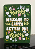 A rectangular dark green decorative card with rounded corners, featuring the phrase “WELCOME TO EARTH LITTLE ONE” in large white letters with yellow outlines, surrounded by hand-painted style flowers, leaves, and small handprints. The sign is photographed leaning on a glossy black surface against a light off-white wall.