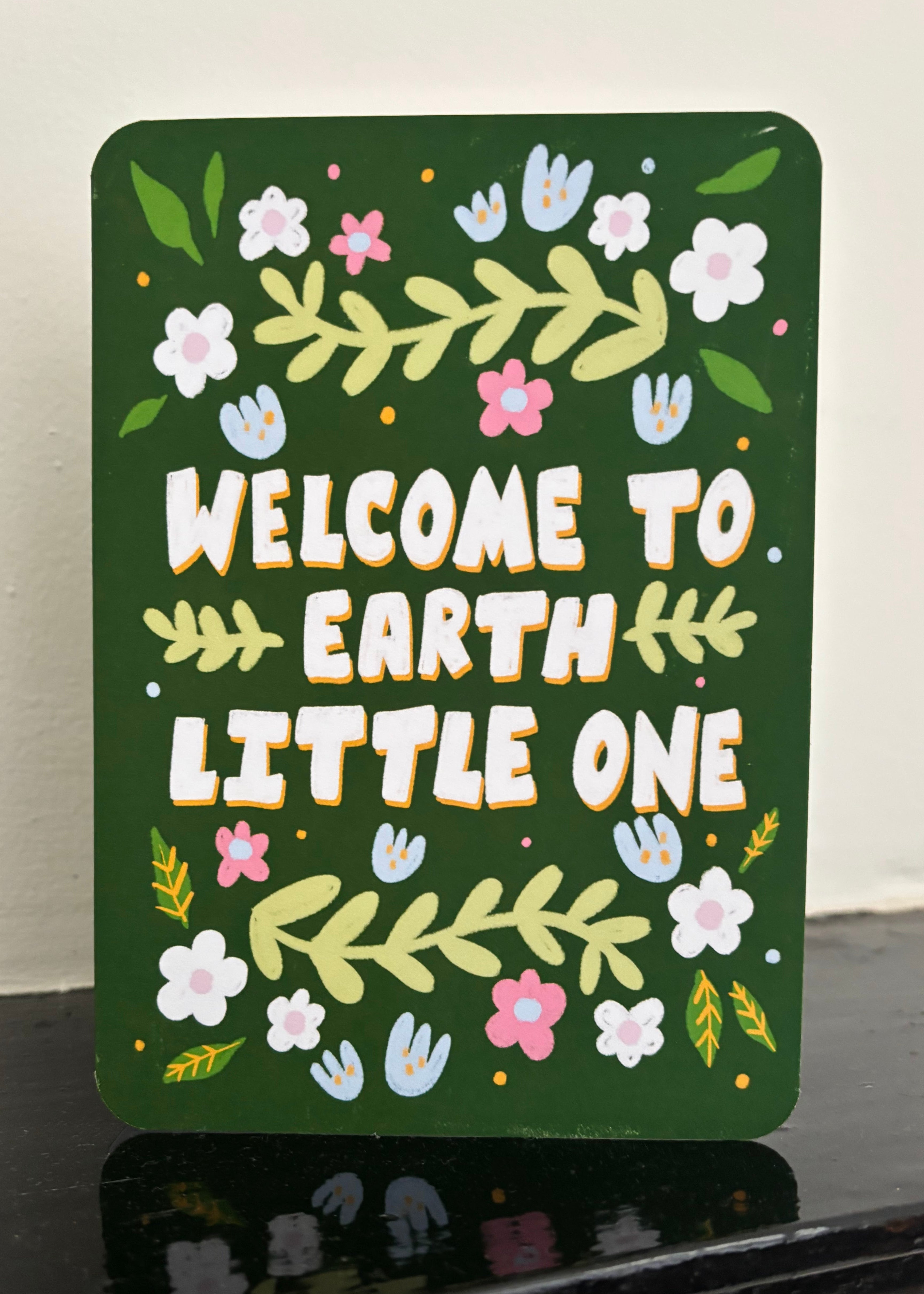 A rectangular dark green decorative card with rounded corners, featuring the phrase “WELCOME TO EARTH LITTLE ONE” in large white letters with yellow outlines, surrounded by hand-painted style flowers, leaves, and small handprints. The sign is photographed leaning on a glossy black surface against a light off-white wall.