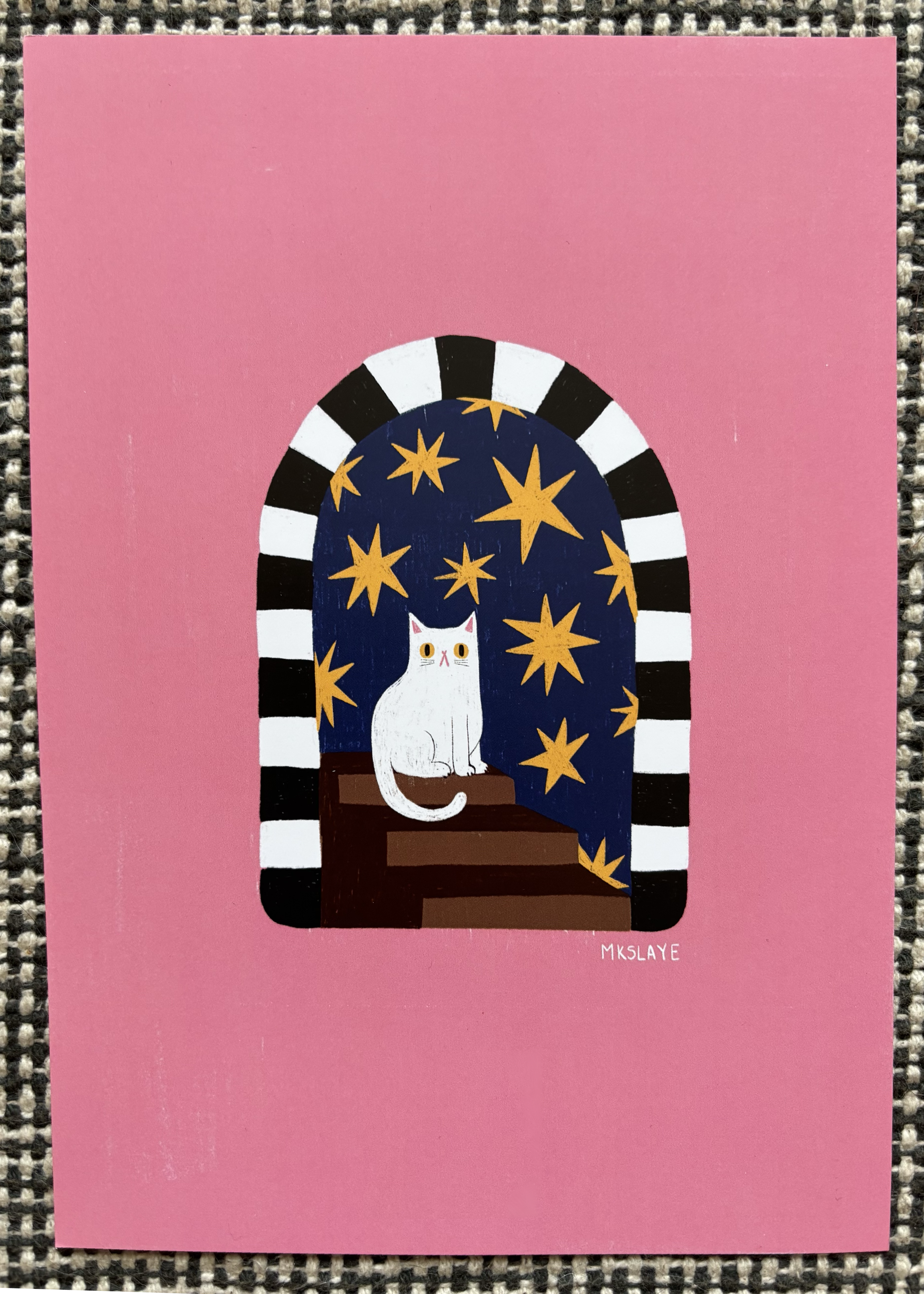 Awhimsical illustration of a white cat sitting on a window ledge under a starry night sky, framed by a black and white striped arch. The card is photographed flat against a textured gray woven background.