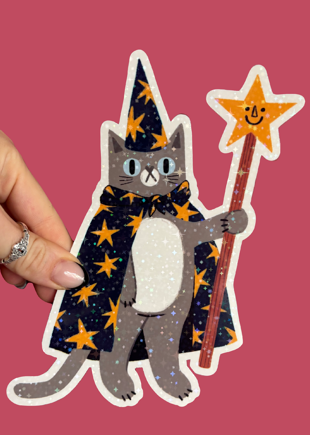 A hand holds a die-cut sticker featuring a gray cat dressed as a wizard with a dark blue starry cape and matching pointed hat, holding a gold star wand, against a solid pink background.