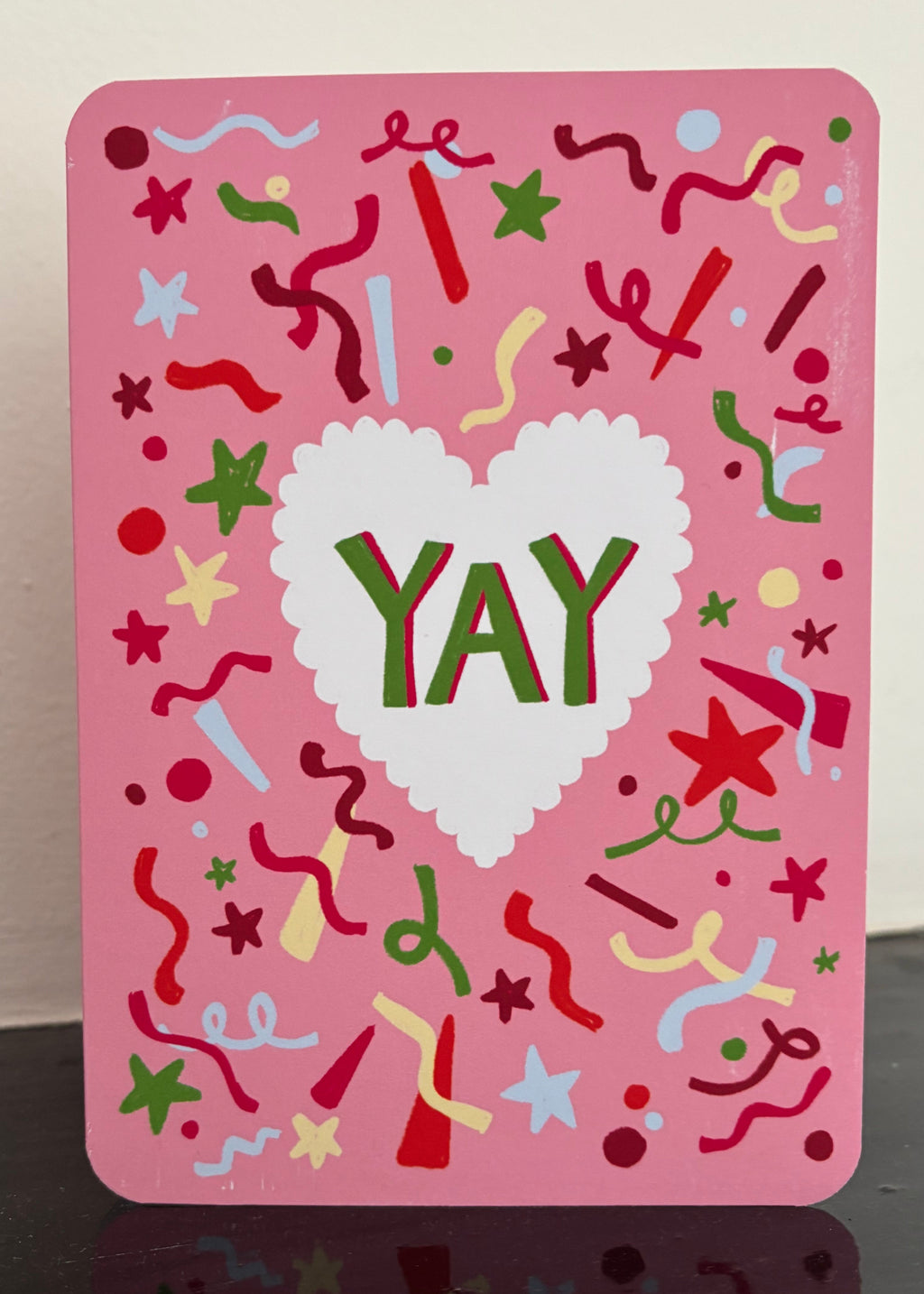 A pink greeting card with a white scalloped heart in the center and the word 'YAY' in green and red. The card is decorated with colorful confetti, stars, and streamers on a pink background.